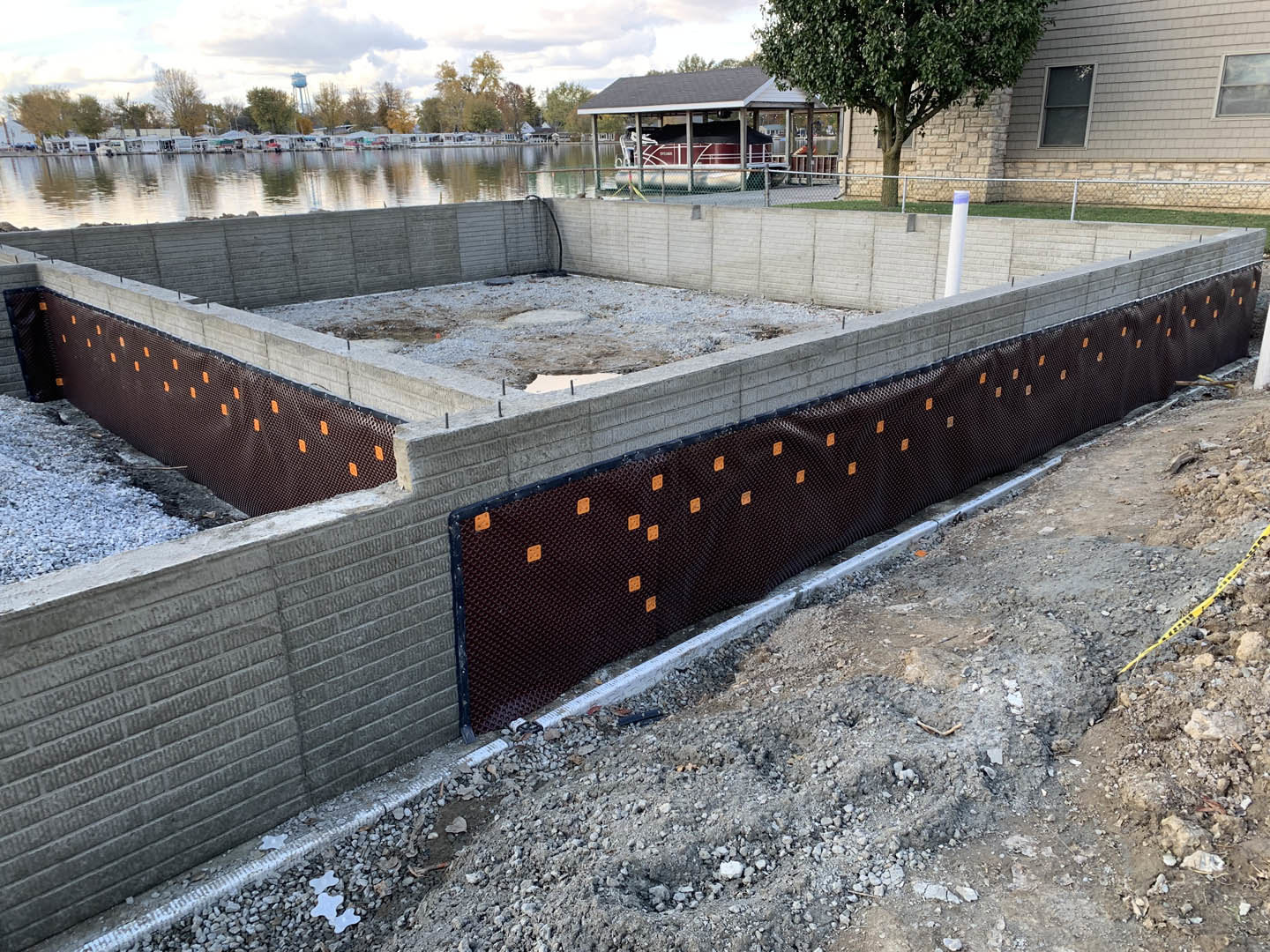 Concrete house foundation surrounded by a wooden fence, tree nearby, covered area with a boat, window visible on adjacent building, waterfront homes in the background.
