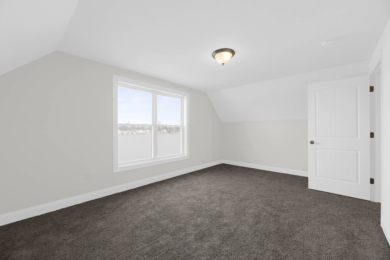 Carpeted room with white plaster walls, white door featuring a silver handle, large window overlooking water and clear sky, modern ceiling light fixture