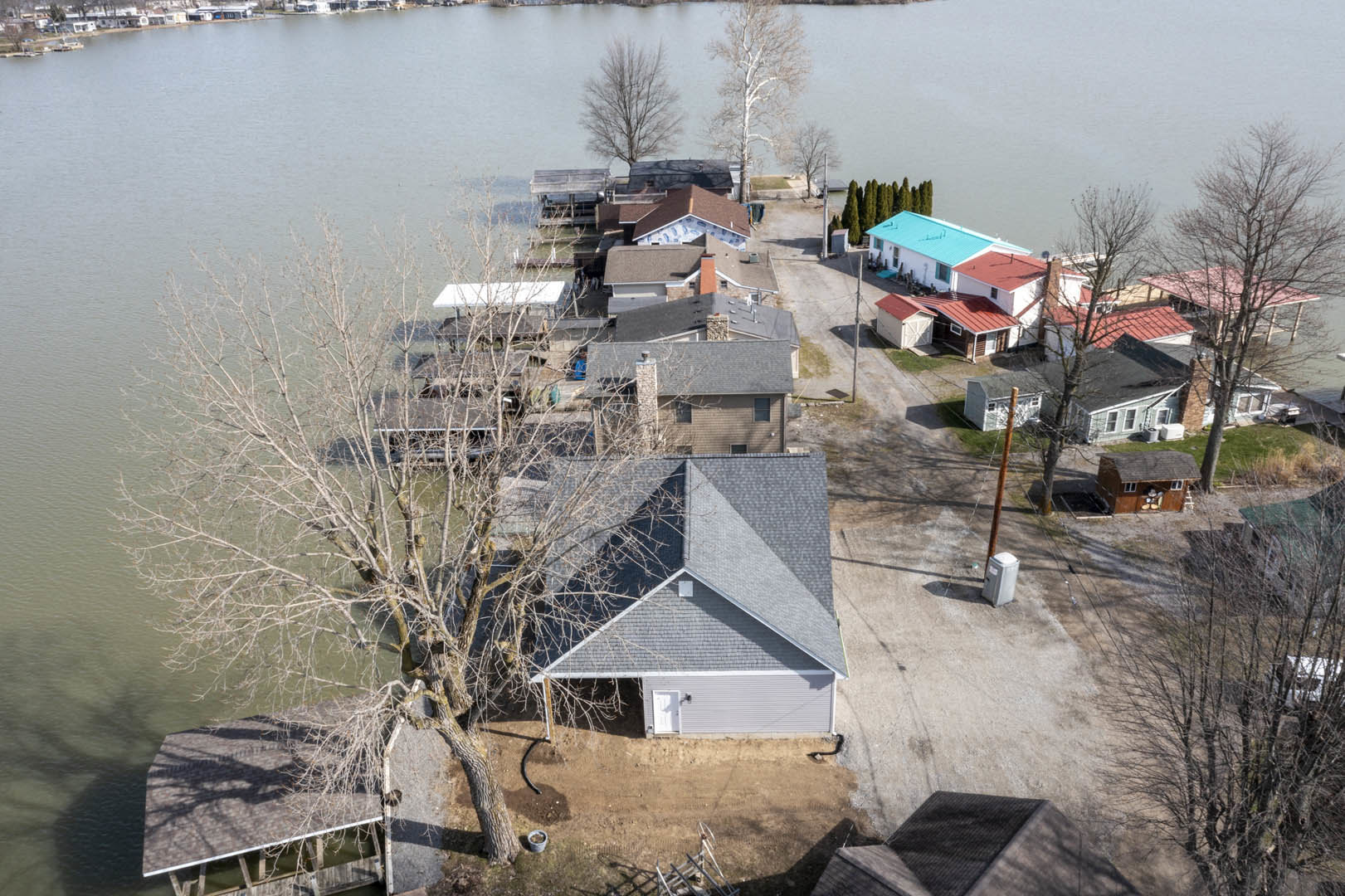 Cluster of lakeside houses with black and gray roofs, surrounded by trees and greenery, white trash can visible near shoreline