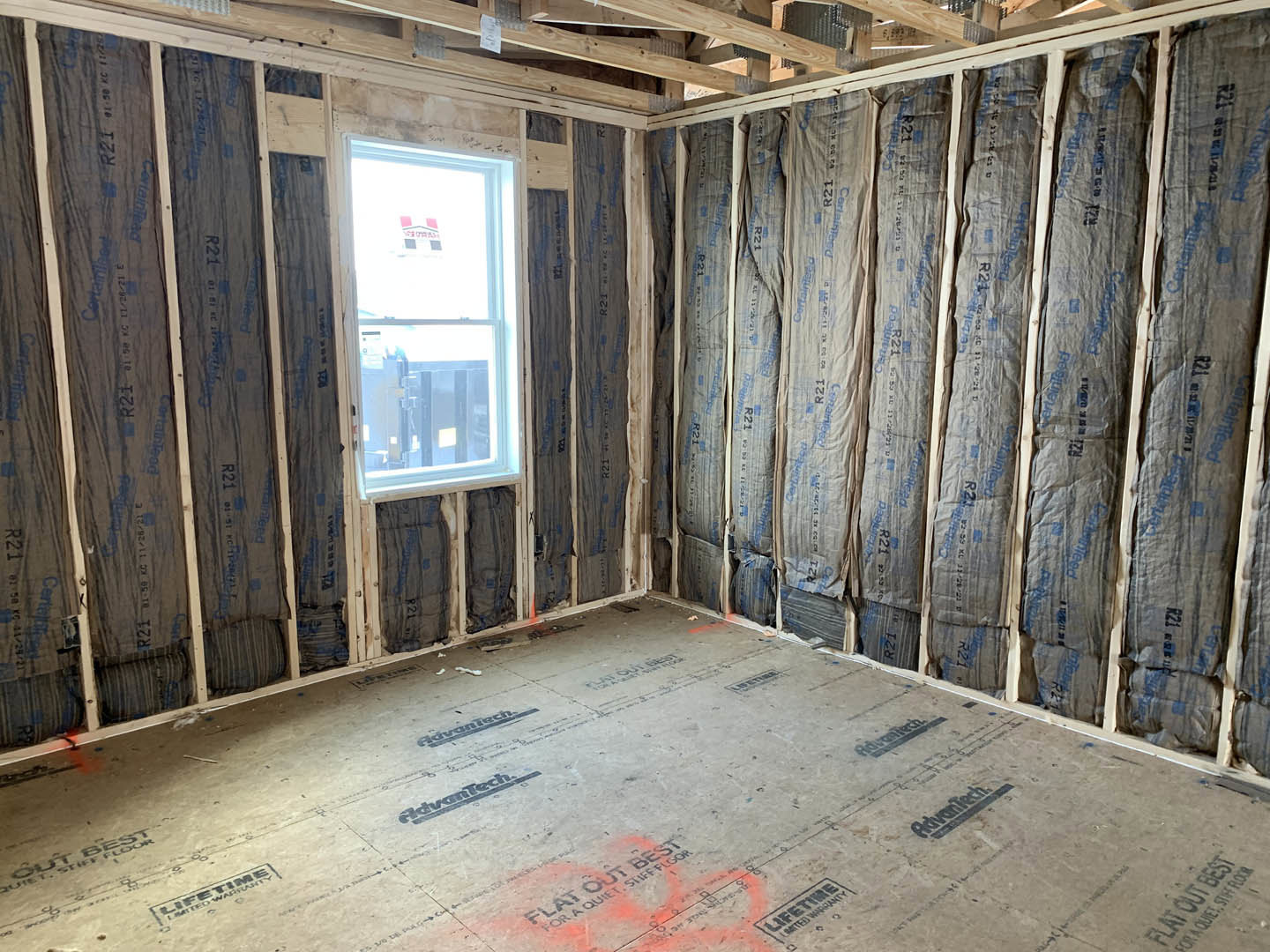 Exposed wood ceiling beams, large window with temporary sign, unfinished walls with insulation, subfloor marked with black writing