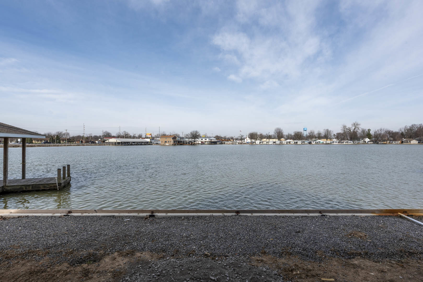 Gravel shoreline leading to a wooden dock on a lake, with custom homes and trees lining the opposite bank under a partly cloudy blue sky