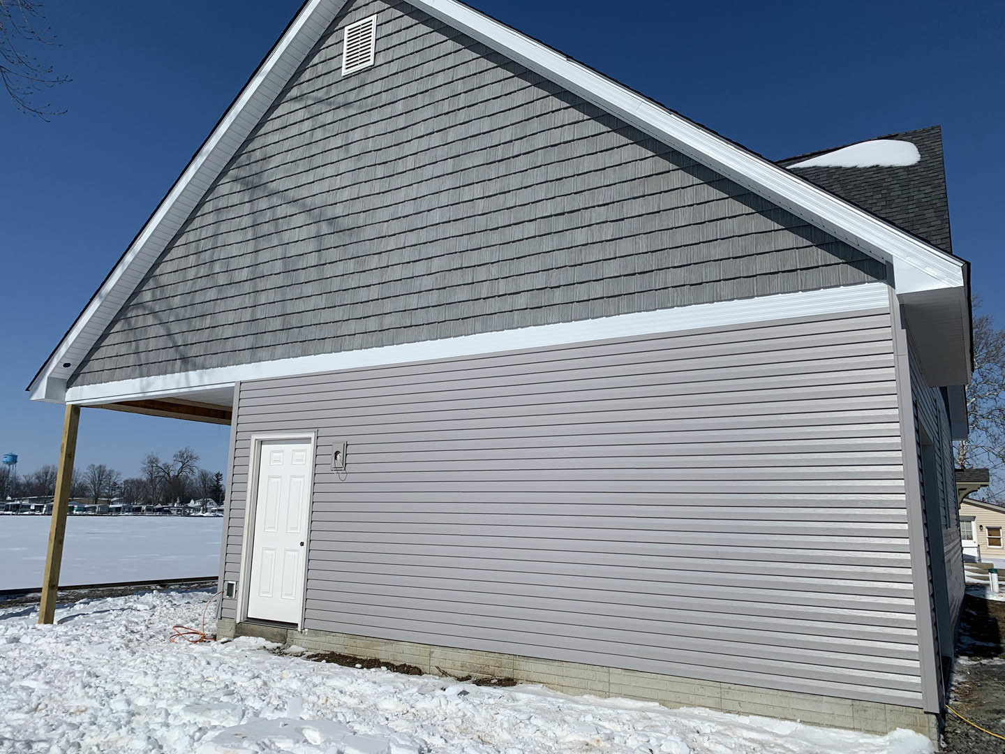 Two-story home with light siding, white front door, attached garage, snow-covered ground, visible roof vent, and wooden utility pole in foreground