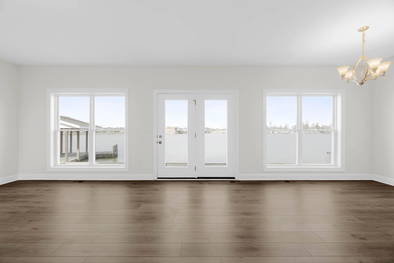 Wood floor and white walls in a bright room, white double doors with glass panes open to a view of a dock and water, chandelier hanging from ceiling.