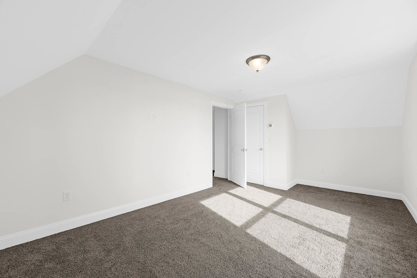 White paneled door with silver handles open to a carpeted room, sunlight streaming onto beige carpet, white walls, and ceiling-mounted light fixture with white shade