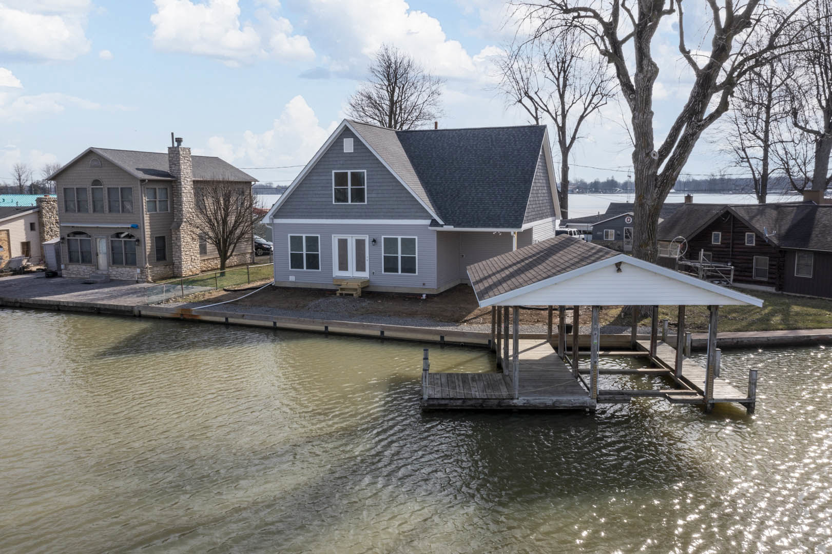 Two-story lakeside house with stone chimney, gray shingle roof, large windows, and a white-roofed boat dock extending over calm water, surrounded by trees under a cloudy sky