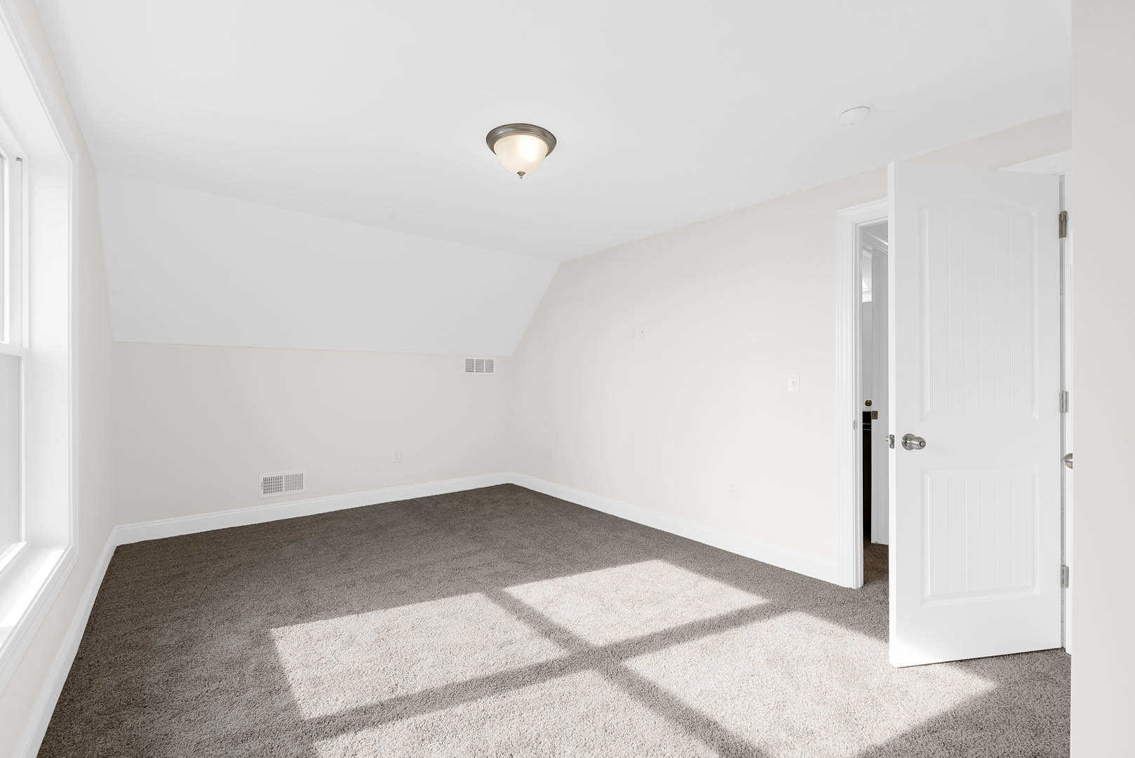 White door with silver knob open to carpeted room, ceiling light fixture illuminated, white walls, window with trim, small cross on floor.