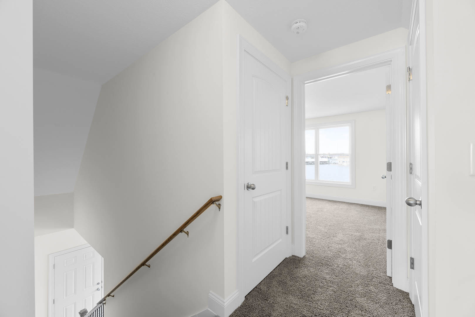Hallway with light-colored flooring, white walls, staircase with wooden handrail, white door, ceiling smoke detector, window overlooking cityscape