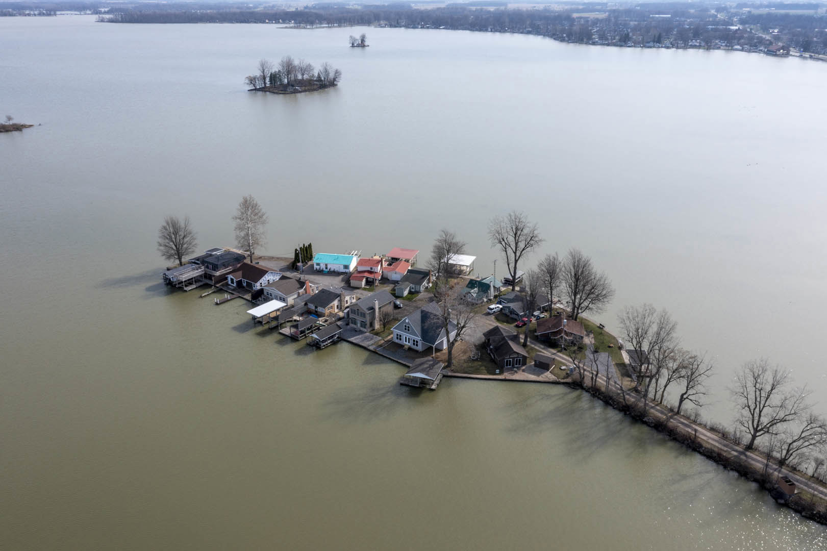 Cluster of houses with blue roofs surrounded by leafy trees on a small island in a lake, calm water reflecting the sky, boats moored along the shoreline.