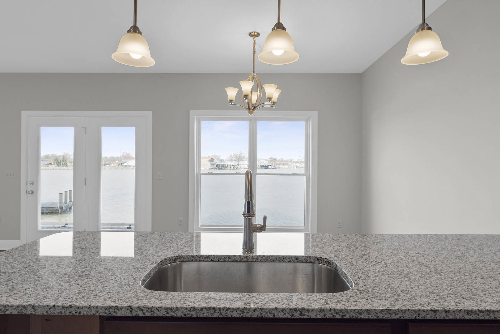 White kitchen with marble countertop, stainless steel sink and faucet beneath large windows, pendant light overhead, glass-paneled door opens to water view