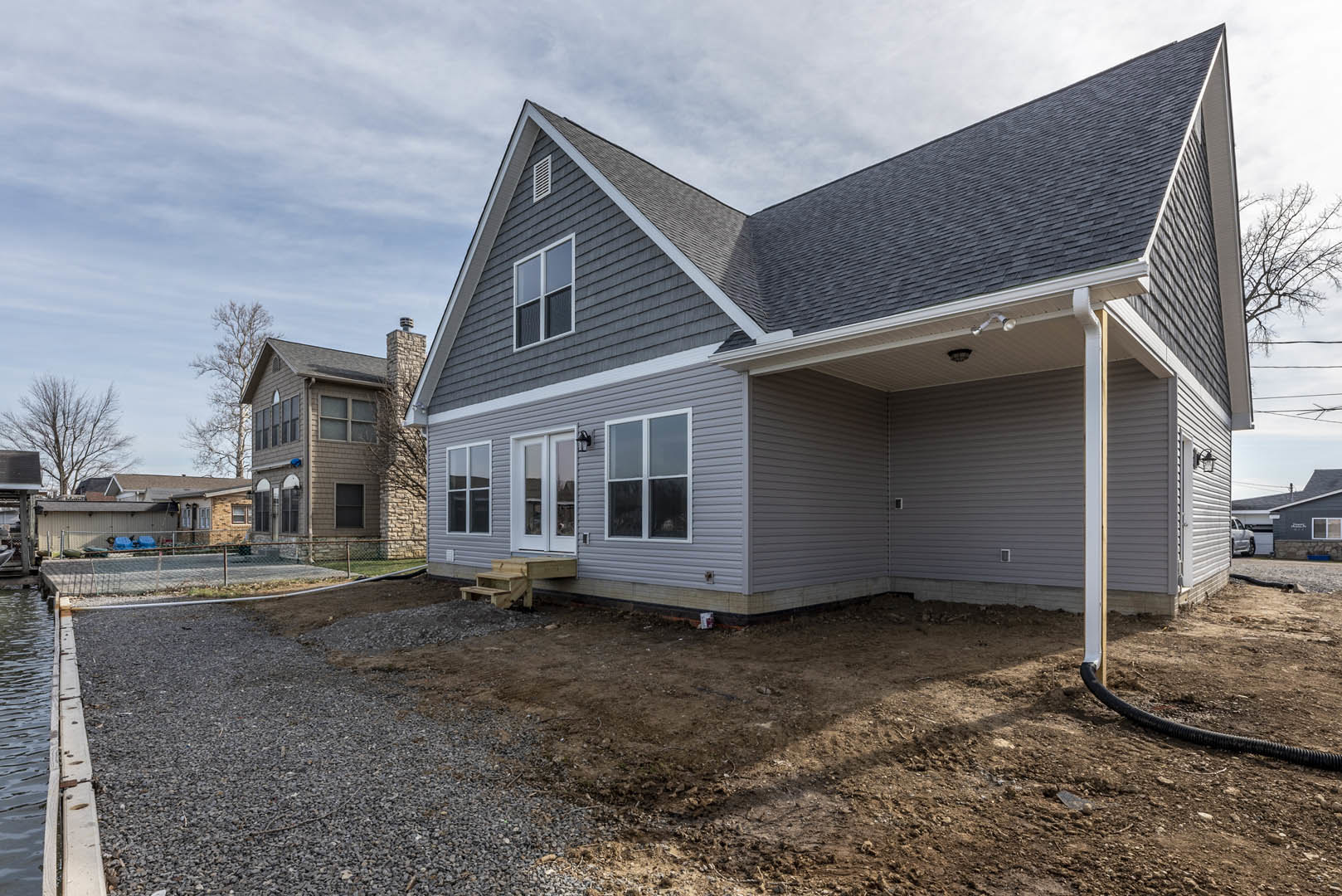 Two-story home with white-framed windows, brick chimney, covered porch, and gravel driveway bordered by a dirt patch under a partly cloudy sky