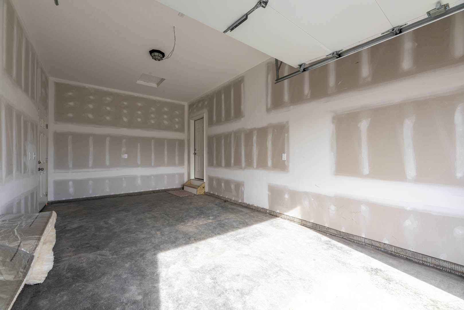 Garage door with silver hardware set in white plaster walls, concrete floor with a step, partial view of staircase, recessed ceiling lighting