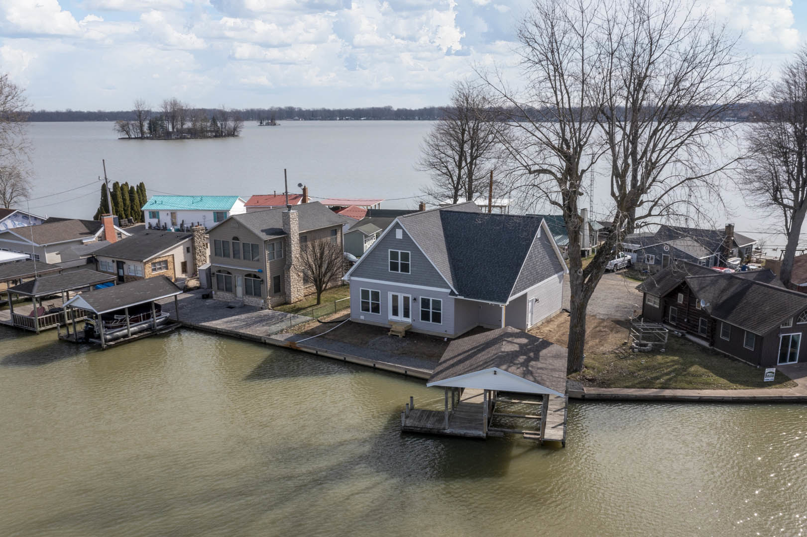 Modern lakeside home with gray metal roof, large windows, and wood siding, surrounded by trees and overlooking calm water under a cloudy sky.