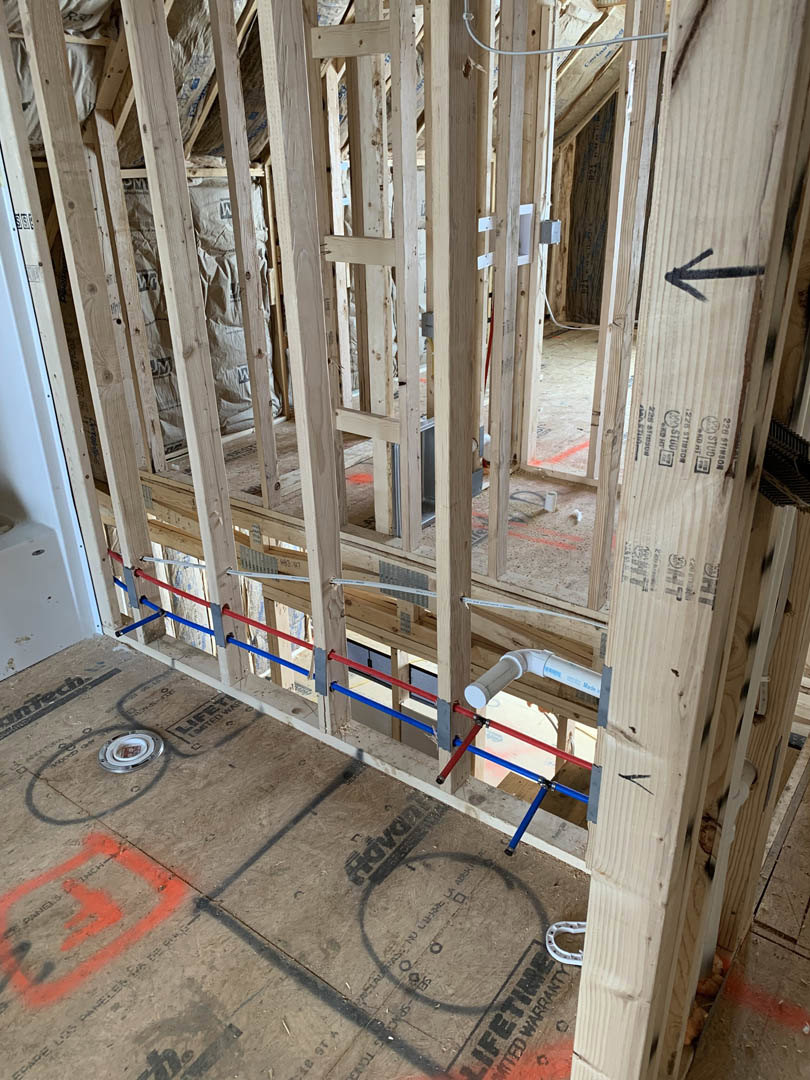 Wood framing with exposed pipes and electrical wires, red and blue plumbing lines running across subfloor, circular drain opening, white horseshoe-shaped object on unfinished