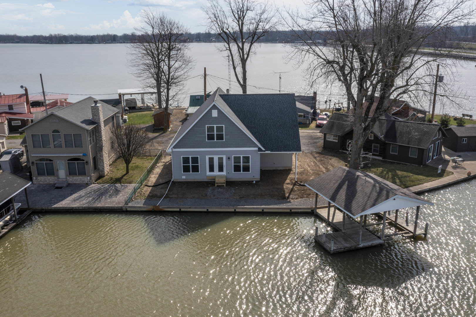 Two-story house with stone chimney and white-framed windows, gray roof, situated beside a river with a boat dock, surrounded by trees and cloudy sky