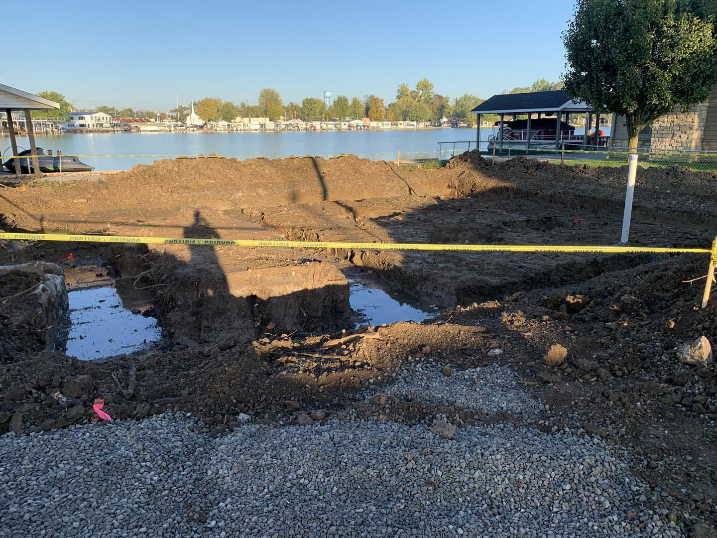 Yellow caution tape stretched across a dirt mound at a residential construction site, tree and partially built home in background