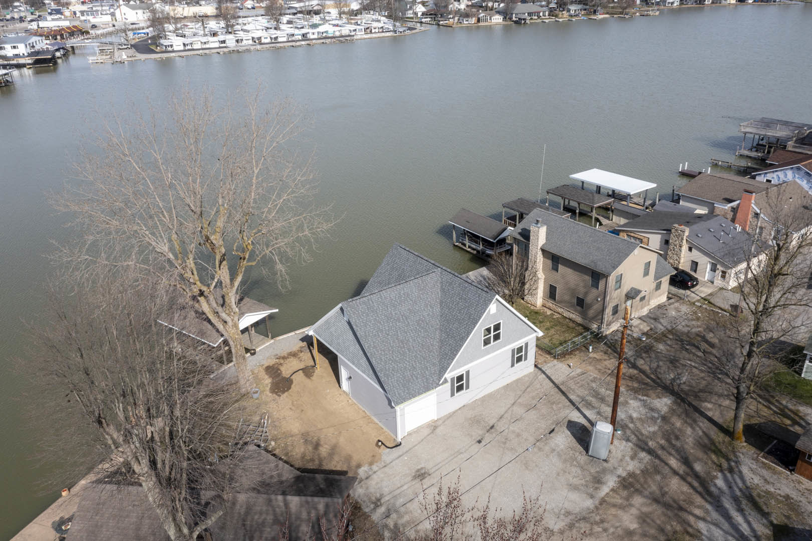 Modern lakeside home with dock, white exterior, large windows, mature tree, white patio table and trash can on lawn, neighboring houses visible across water.