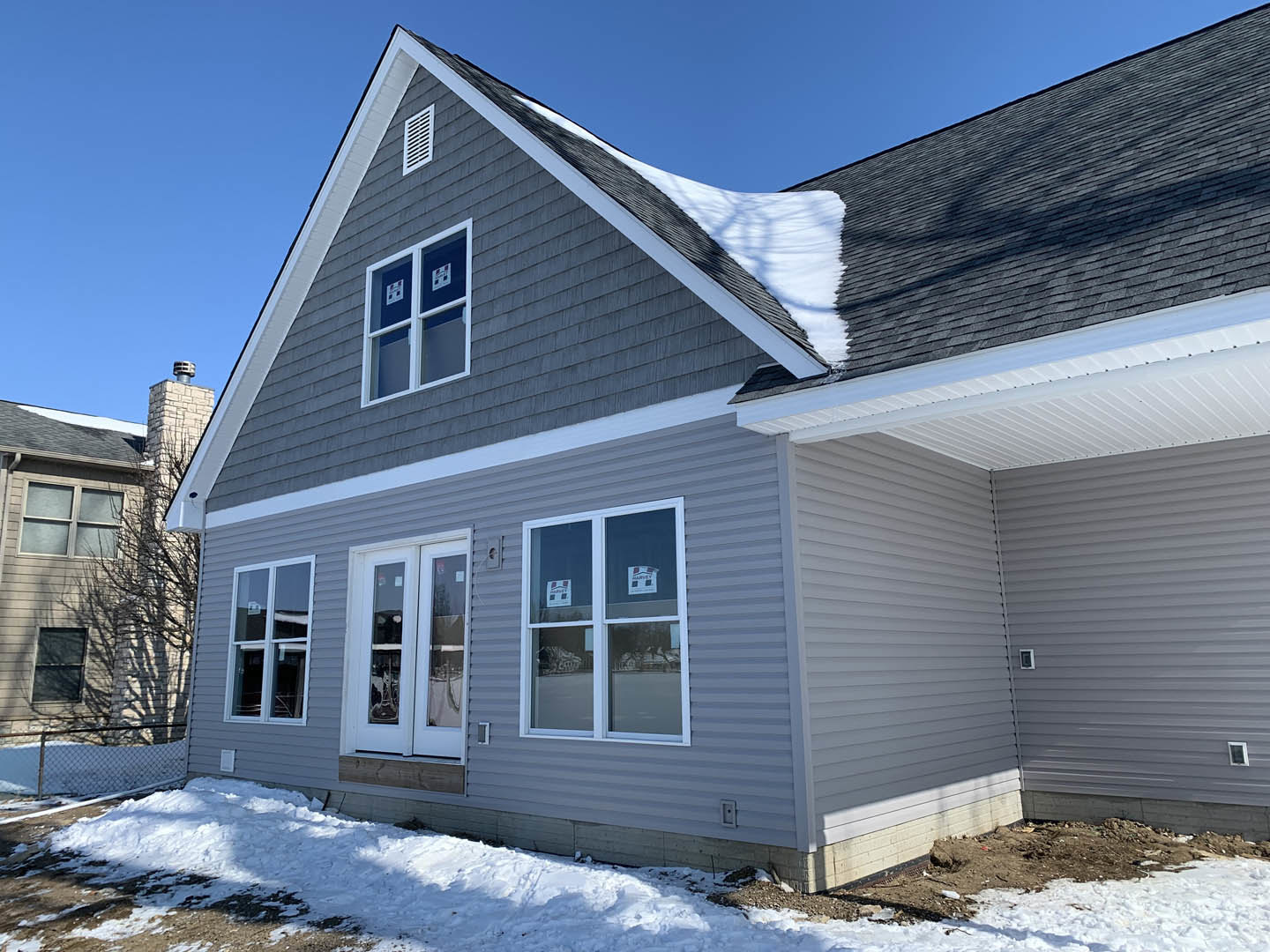Two-story house with gray siding, snow covering the roof and ground, large windows with stickers and metal bar, overcast winter sky