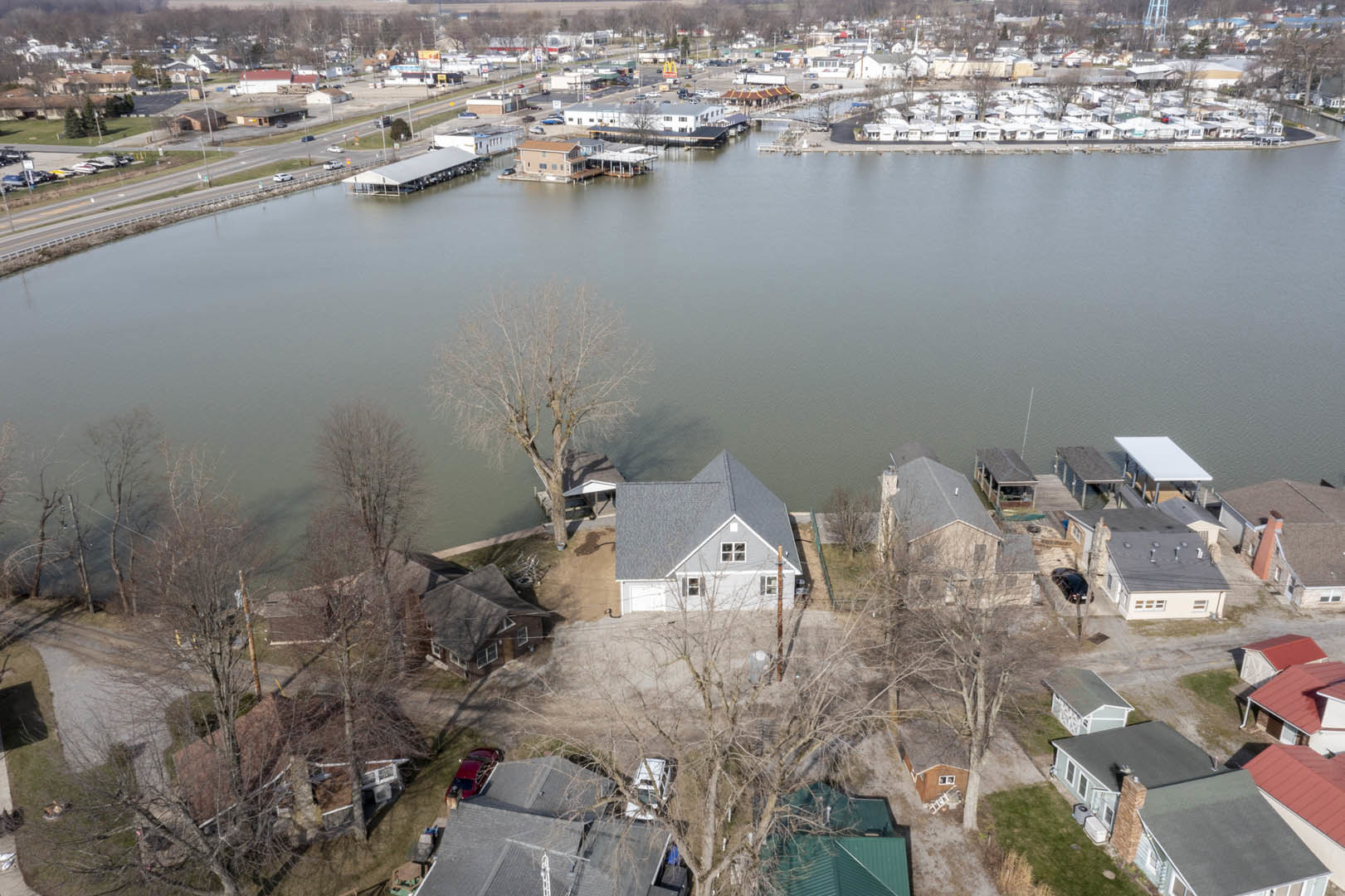 Aerial view of a city beside a river, houses with triangular roofs lining the waterfront, boats docked along the channel, trees scattered near the shoreline