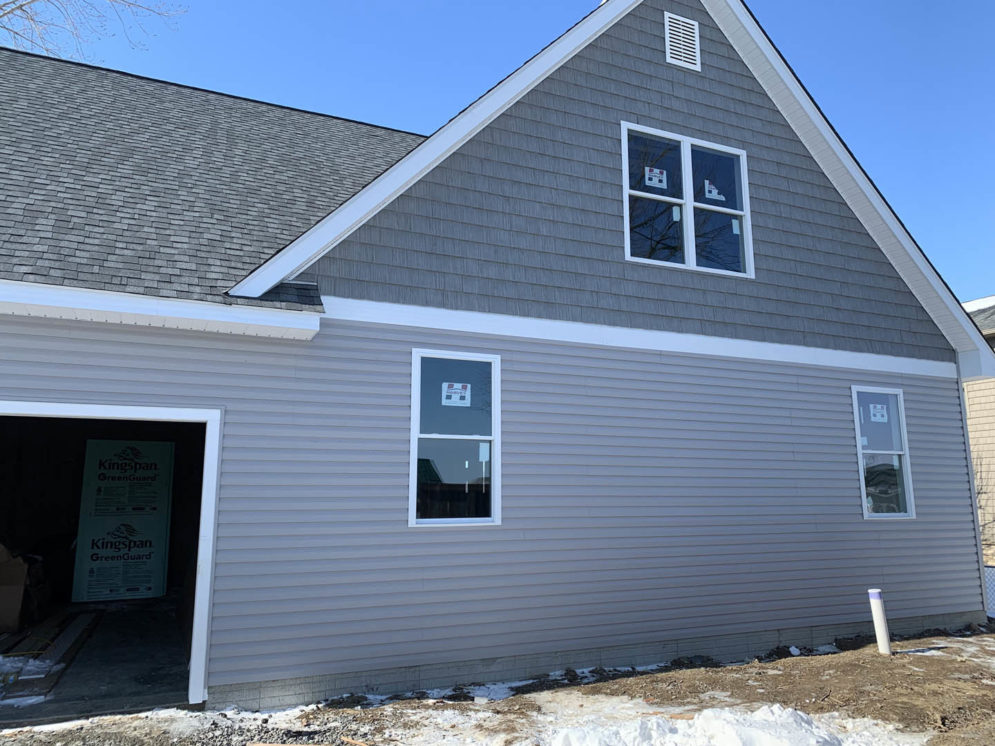 Light gray siding custom home with attached garage, open front door, white-framed windows featuring stickers and signage, gable roof, and paved driveway under clear sky.