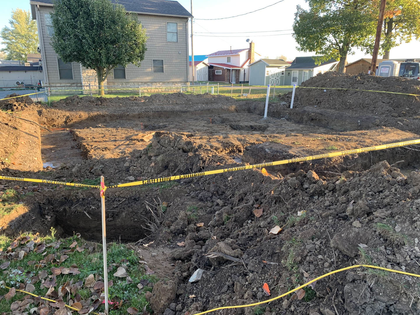 Dirt field bordered by yellow caution tape, tree in front of house with red roof, white rock on ground, garden area with mulch and compost