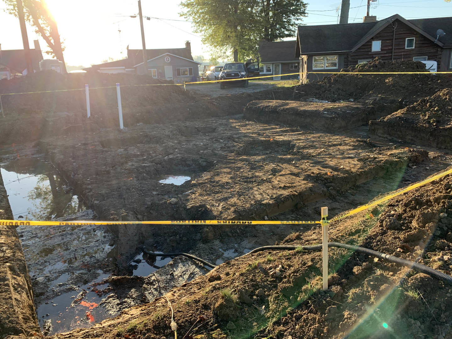 Log cabin under construction with yellow caution tape surrounding a dirt field, black truck parked nearby, green-leafed tree, and basketball hoop on a metal pole.