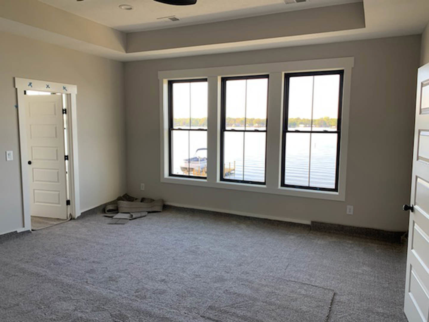Carpeted room with large windows overlooking water, white door with black hardware, light grey walls, natural daylight.