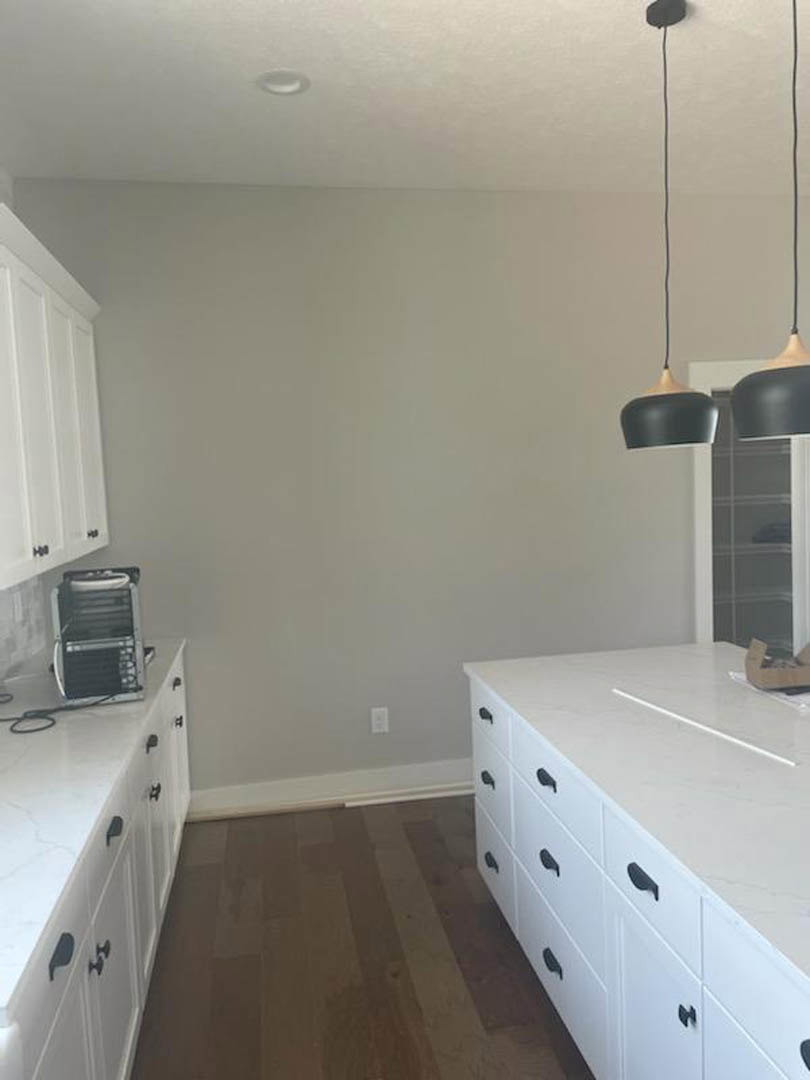 White kitchen cabinets with black pendant lamps, white countertops with black handles, wood flooring, and a kitchen cart near a white door.