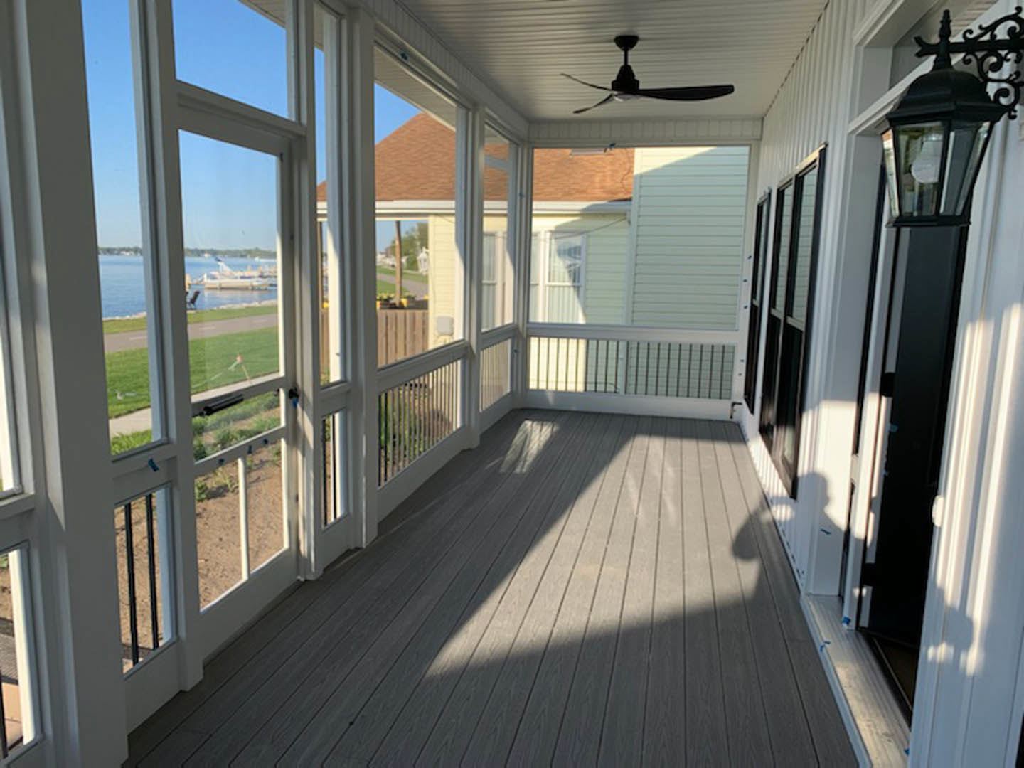 Covered porch with wood plank flooring, ceiling fan, white railings, glass-paneled door, and large window overlooking outdoor area