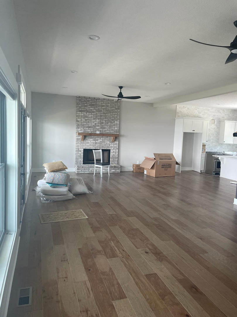 Living room with laminate wood flooring, white upholstered chair with black cushion, carpet detail, brown storage box, built-in fireplace with white mantel shelf, plaster walls