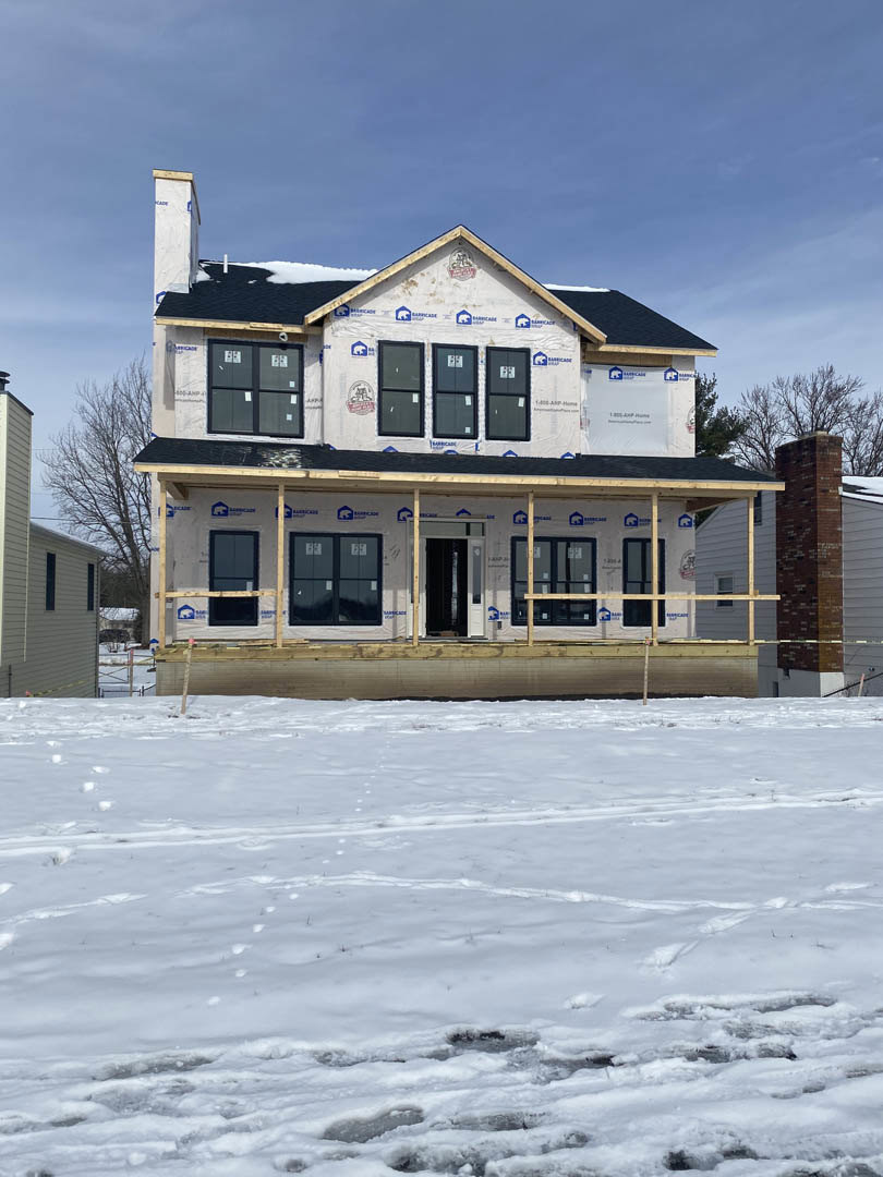 Partially built brick house with exposed framing, snow-covered ground and footprints, wooden deck, brick chimney, windows with construction signs, close-up of unfinished door
