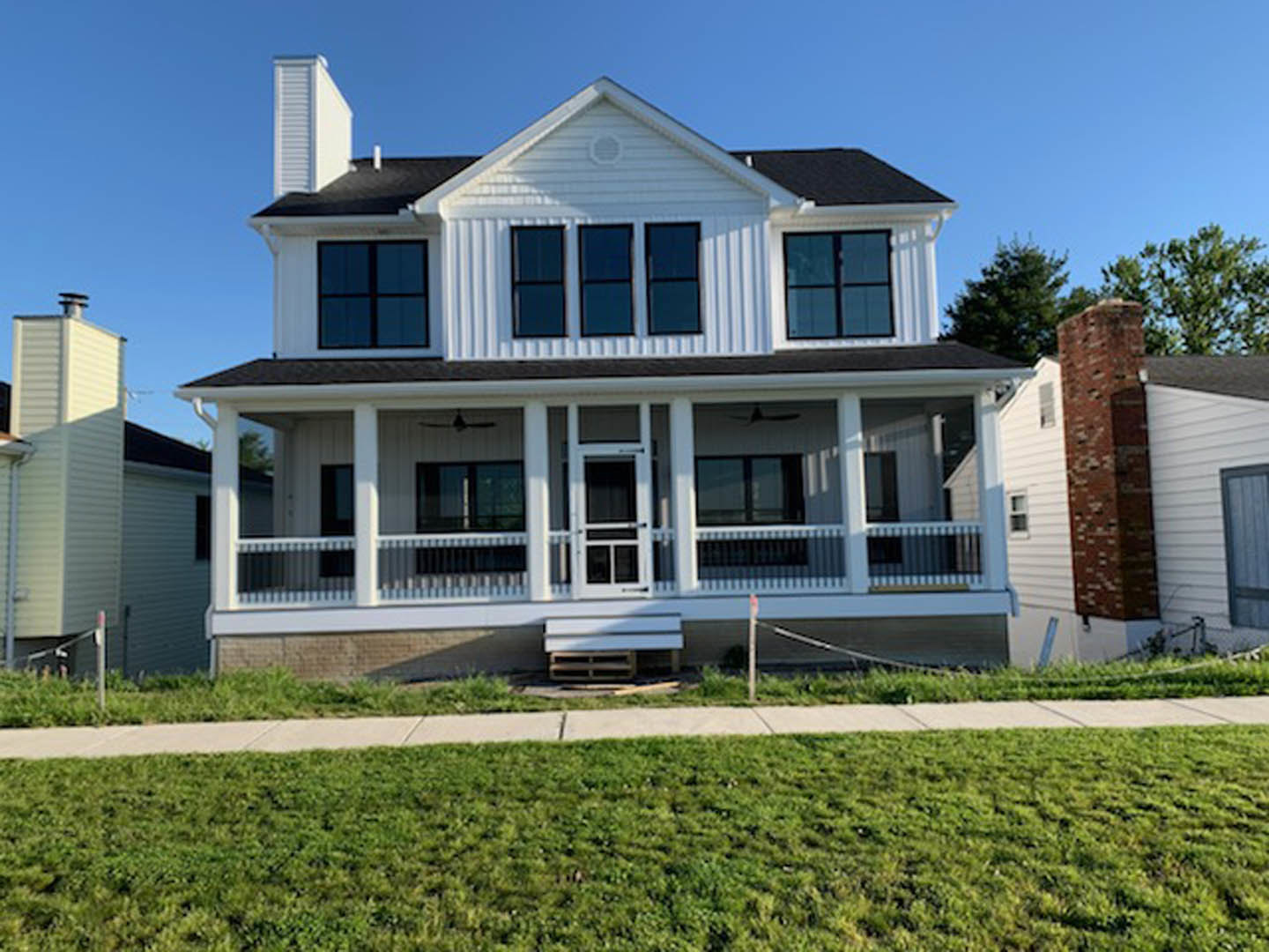 White two-story house with covered porch, black screened door, brick retaining wall, green lawn, and sidewalk in front.