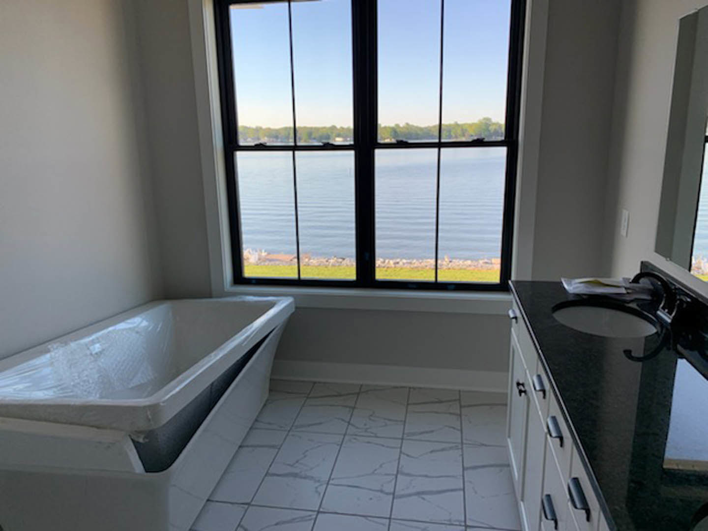 White freestanding bathtub wrapped in protective plastic, large window with outdoor view, light gray tile floor, chrome faucet, white walls
