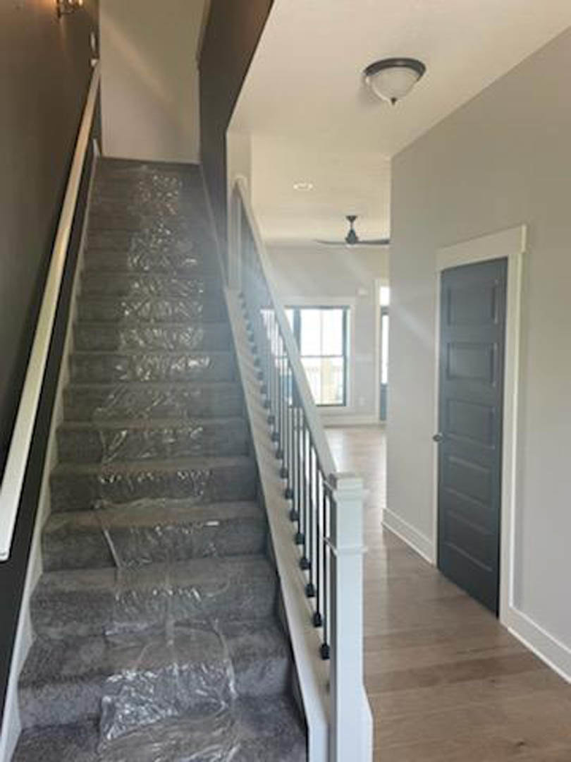 Wood staircase wrapped in protective plastic, white walls, metal handrail, light fixture overhead, window letting in natural light