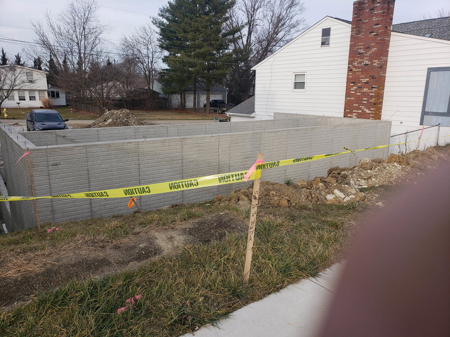 Fenced yard with green grass, large tree, brick chimney, white exterior wall, black car parked nearby, yellow caution tape on wooden post