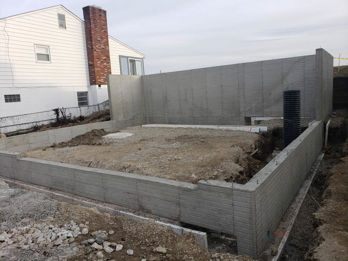 Concrete foundation with exposed dirt and rocks, brick chimney rising above, black pipe with white cap, white-framed window set into exterior wall, cloudy sky overhead