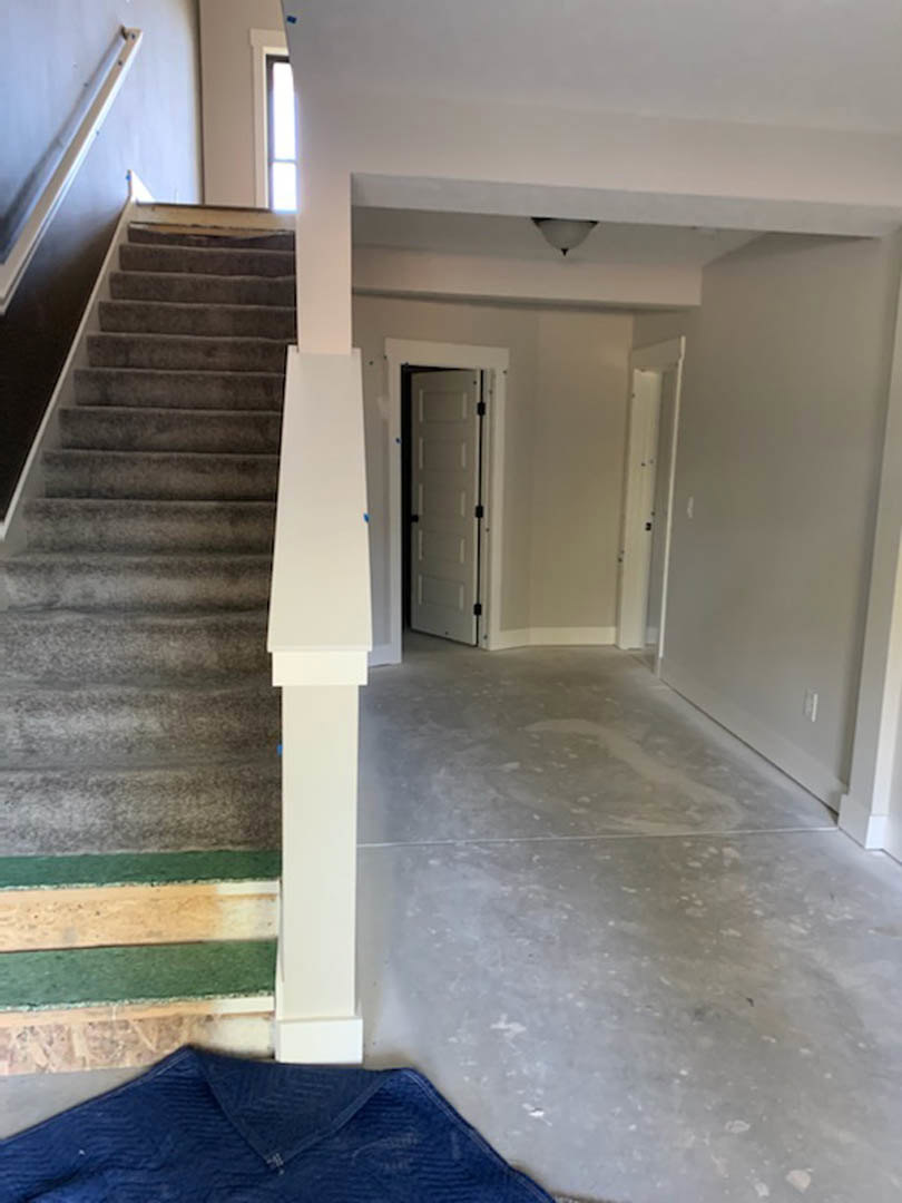 White-painted staircase with wooden treads, black metal railing, white paneled door with black handles, light-colored flooring, blue area rug, white walls