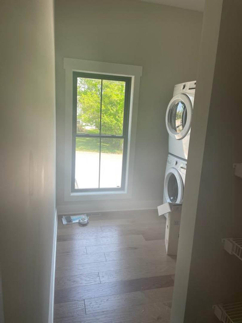 Front-loading washing machine beside a white wall and door, wooden floor, large window with green tree visible outside, ceiling-mounted white square fixture.