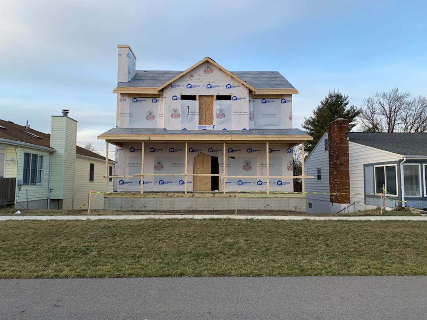 Two-story house under construction with exposed framing, white sheeting, and brick walls, surrounded by a wooden fence and green grass lawn