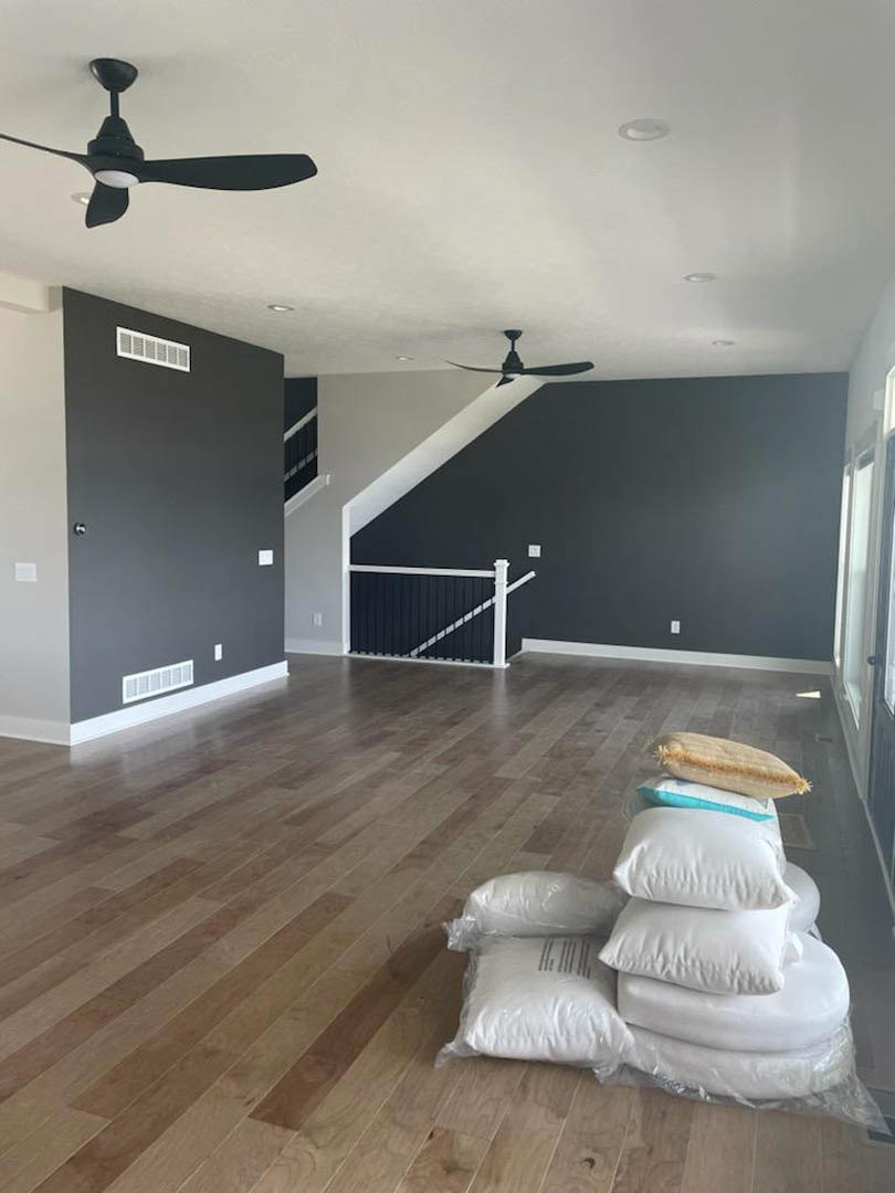 Living room with wood flooring, white couch topped with stacked pillows, ceiling fan with light fixture, plaster walls, and a stack of bags near the gate.
