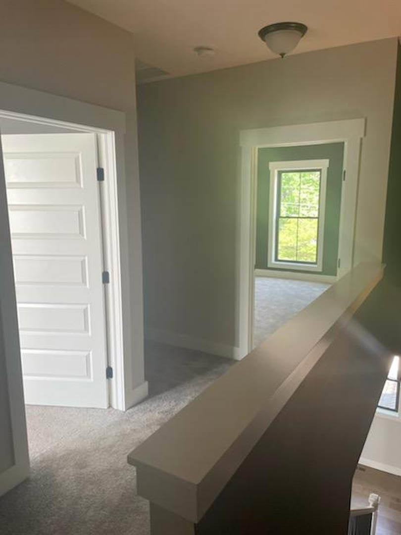 Bright room with light wood flooring, white walls, large window overlooking greenery, white door with black handle, modern ceiling light fixture, and staircase with wood steps and
