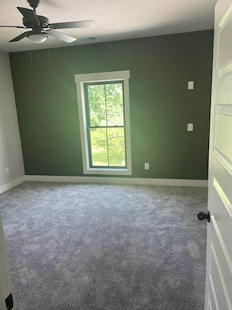 Carpeted bedroom with white walls, ceiling fan, large window overlooking trees, and white door
