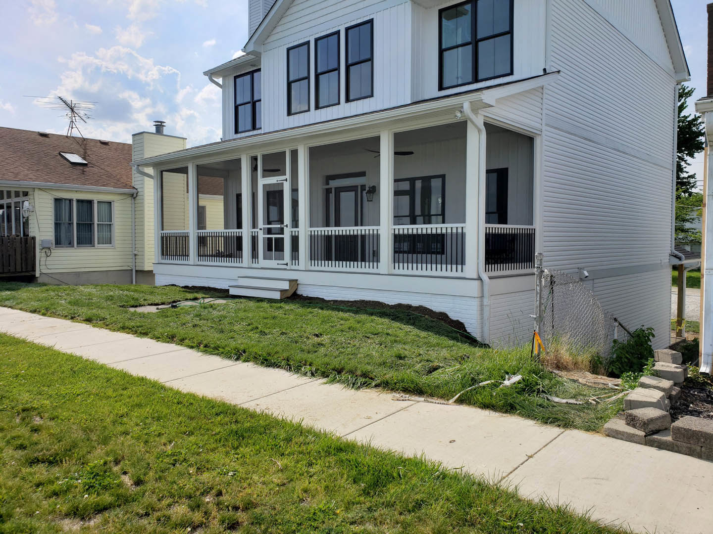 White house with covered porch, multiple windows, grassy lawn, concrete sidewalk, rooftop antenna, blue sky
