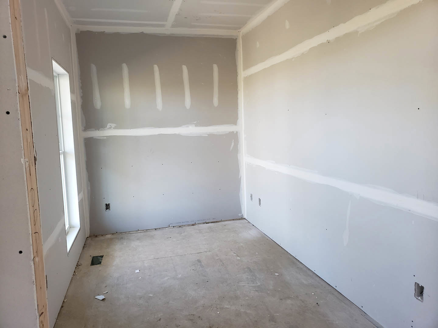 Room with smooth white plaster walls, rectangular window framed in white, light wood flooring, and visible wall holes for fixtures.