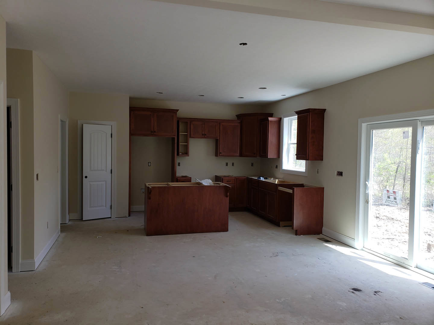 Kitchen with natural wood cabinets, white plaster ceiling, white door with black knobs, window displaying a sign, concrete floor with wood inset, wooden table featuring a hole