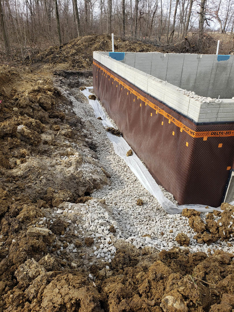 Concrete foundation surrounded by piles of dirt and rocks, bare trees in the background, construction fencing visible along site perimeter
