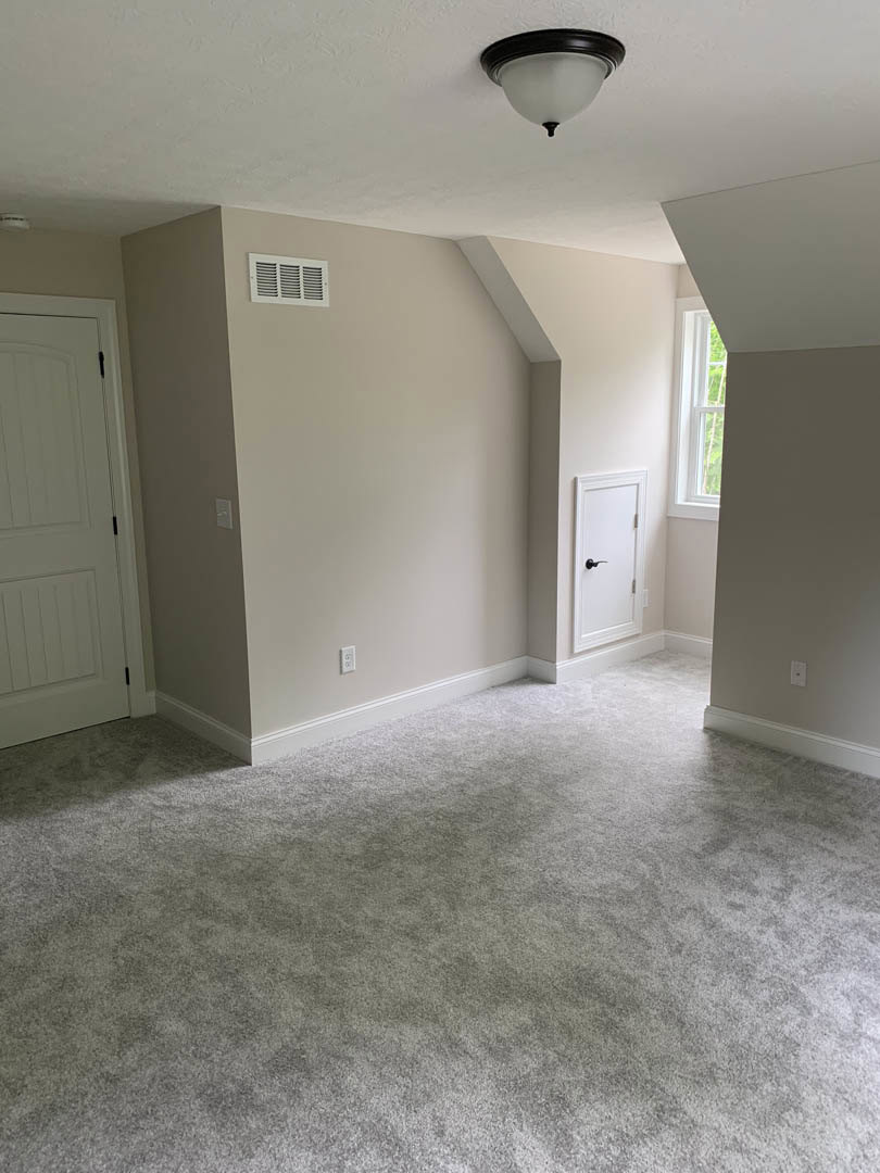 Carpeted room with white walls, white door featuring black handle and hinges, ceiling light fixture, window, and close-up of a vent