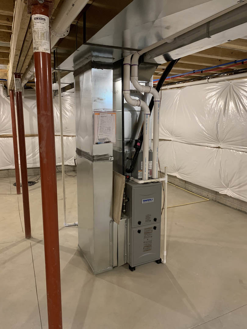 Large silver metal utility box with exposed pipes mounted on a white wall, paper taped nearby, white plastic bag hanging, and black tape on a rectangular object.