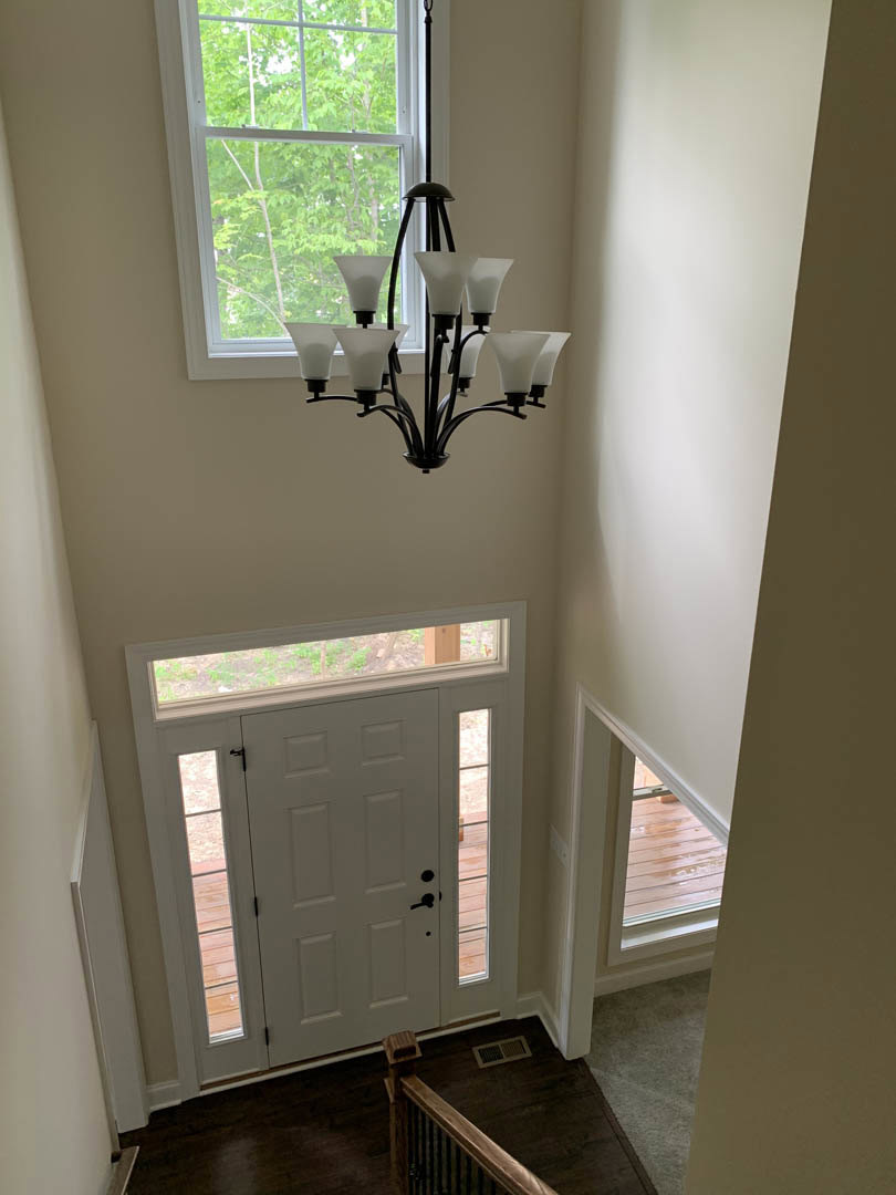 Hallway with white paneled door, glass window showing tree outside, chandelier with white shades, wooden railing, floor vent, and plaster walls