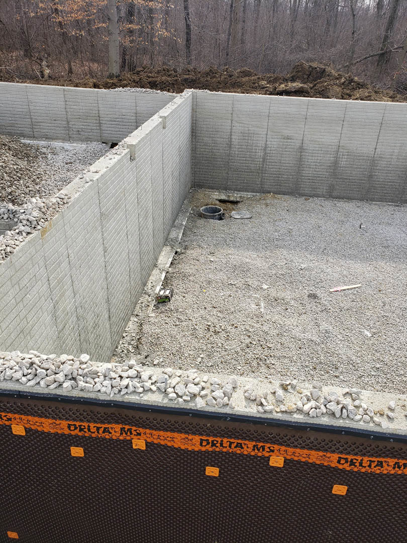 Concrete foundation surrounded by gravel and rocks, adjacent to a dirt pile and trees, at an outdoor construction site