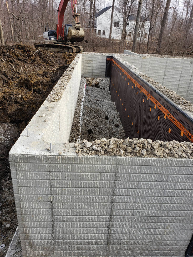 Bulldozer parked beside partially built brick and concrete foundation wall, gravel and dirt piles in foreground, wooded area in background